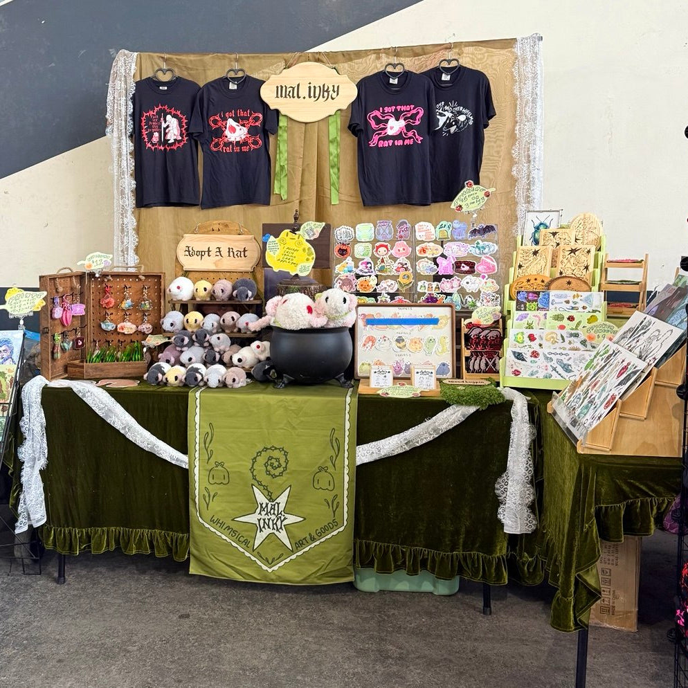 A market stall display featuring gothic-themed merchandise, including three black graphic t-shirts, a green velvet draped table with a central banner, plush toys, stickers, and art cards against a painted concrete wall.